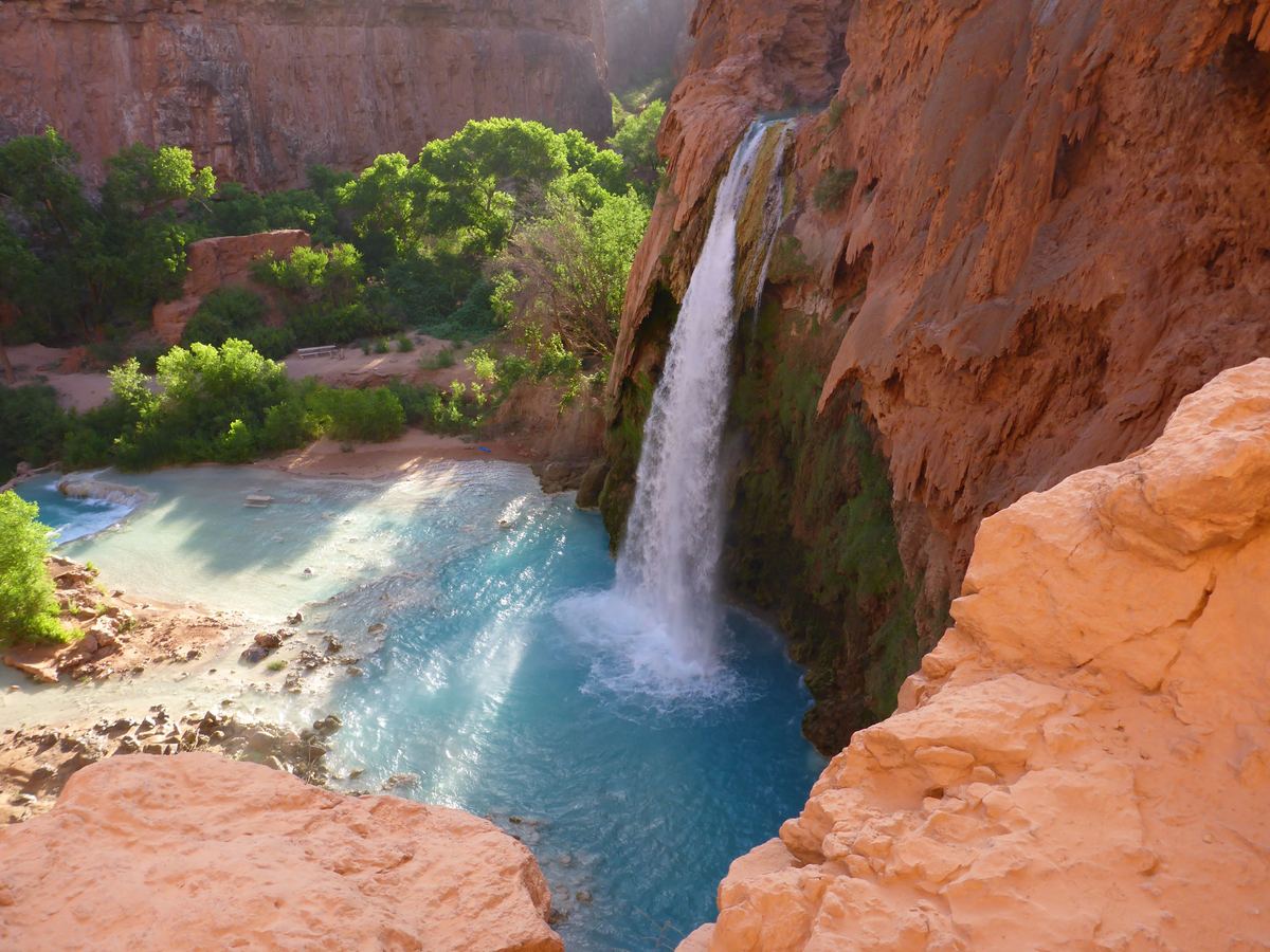A waterfall pours down a red rock cliff into a bright turquoise pool, with green trees and sunlit orange sandstone ledges around it.