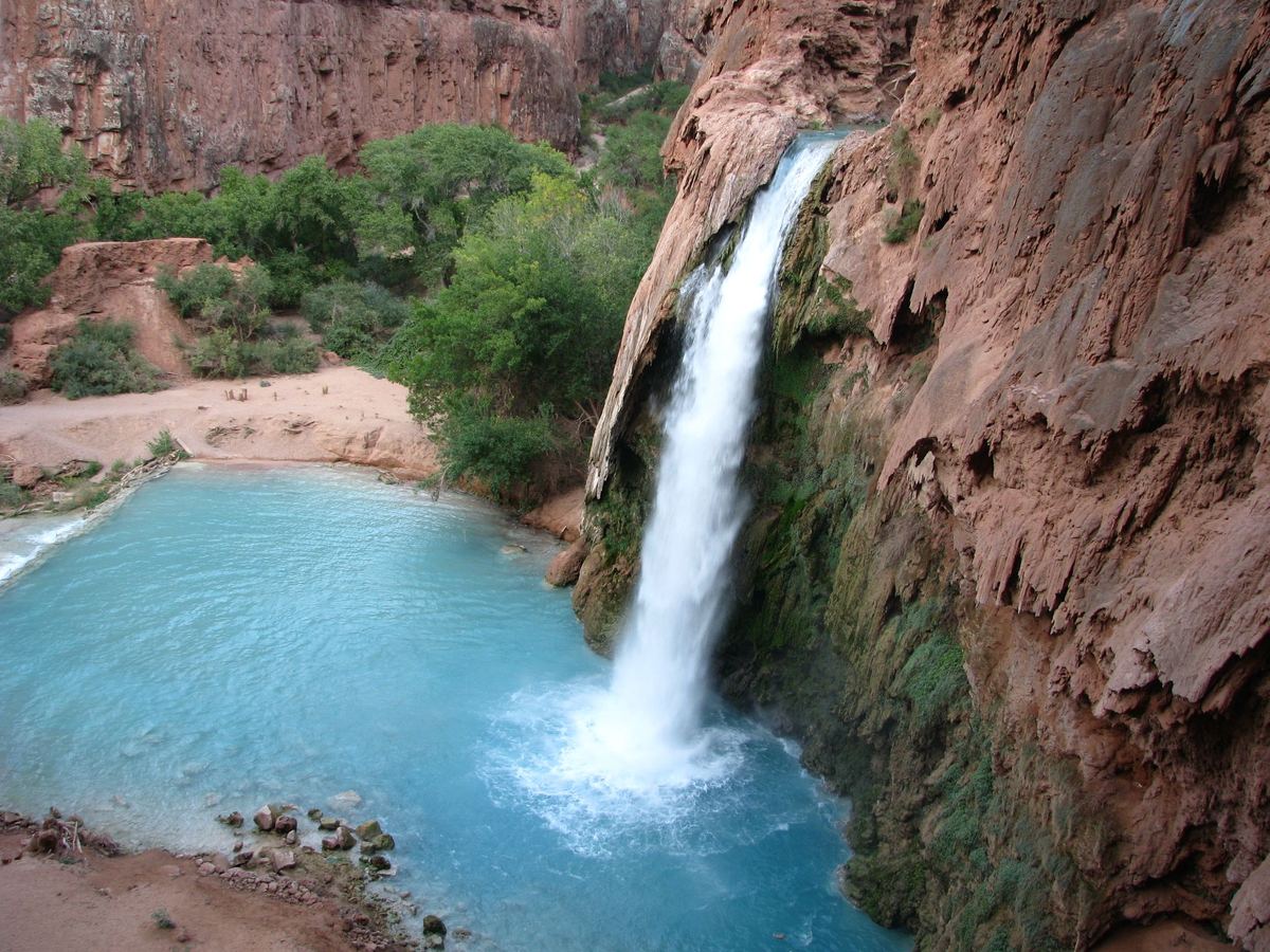 A white waterfall plunges down a red rock cliff into a bright turquoise pool, with green trees and a sandy bank beside it.
