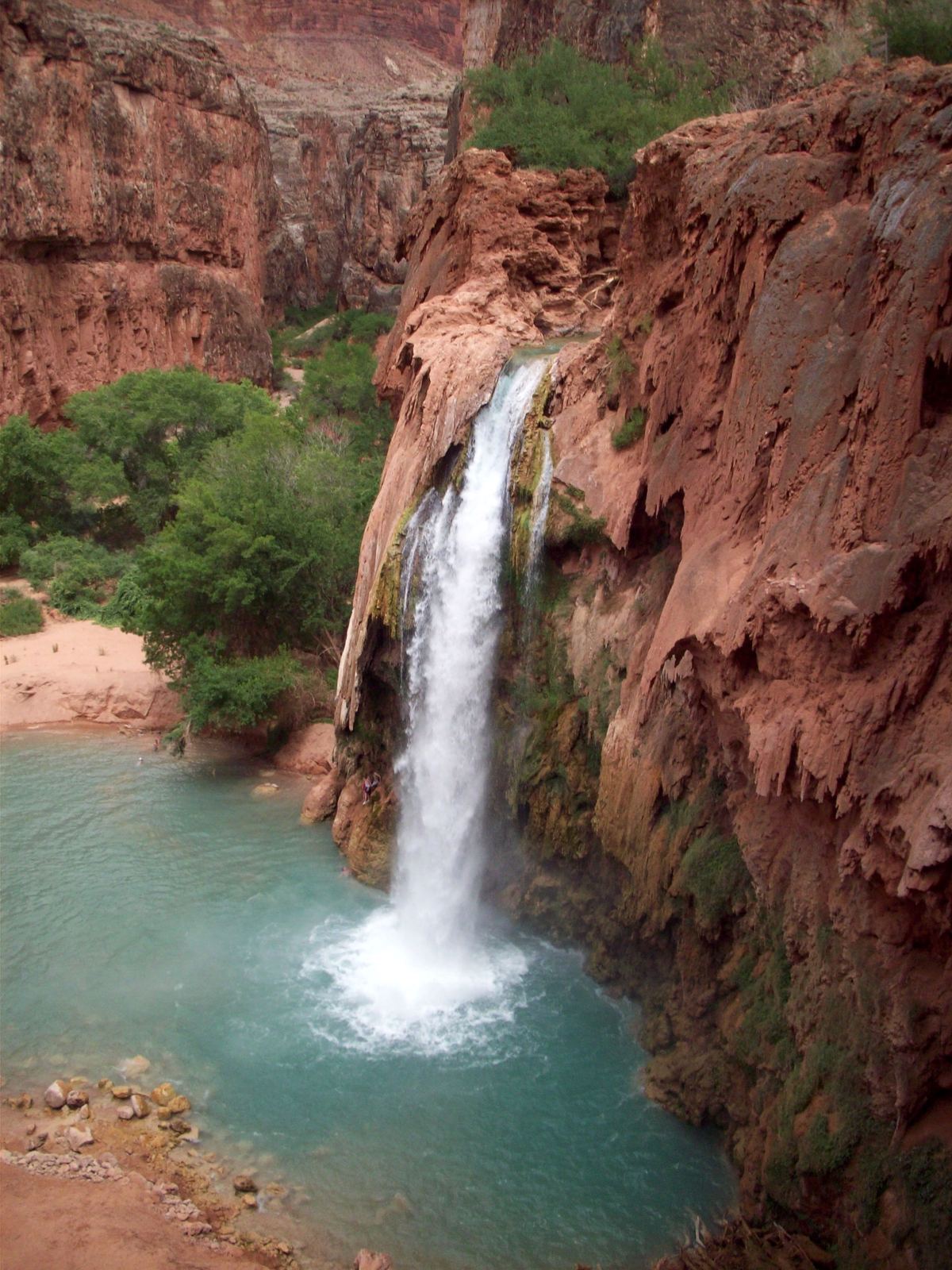 A waterfall plunges down a red rock cliff into a turquoise pool, with green trees on the left and a sandy bank beside the water.